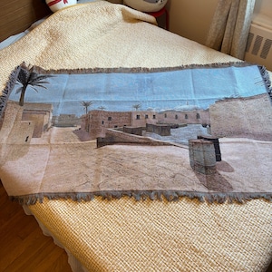 May include: A rectangular woven throw blanket featuring a detailed desert landscape scene with tan buildings, palm trees, and a cylindrical barrel in the foreground. The blanket has a fringed edge and displays a muted color palette of beige, brown, and light blue.