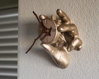 May include: A wall-mounted clock featuring a metallic gold-colored hand sculpture holding a circular clock face. The clock has dark brown, branch-shaped hands and a smooth, round dial. The text kamdes3d.etsy.com is visible at the bottom.