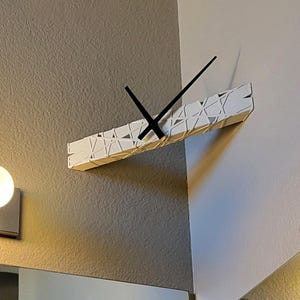May include: A modern, white wall clock with a geometric design. The clock has a rectangular shape with a textured surface and black hands. The clock is mounted on a gray wall, adding a contemporary touch to any space.