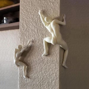 May include: Two off-white, smooth-surfaced wall sculptures depicting human figures in climbing poses. One figure is positioned on the flat surface of the wall, while the other figure is mounted on the corner edge, appearing to scale the vertical structure.