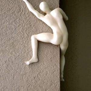 May include: A matte off-white figurine of a human figure in a climbing pose, mounted on a corner edge. The figure has one arm extended upward and one leg bent, with the body positioned to appear as if it is scaling the vertical surface of the wall.