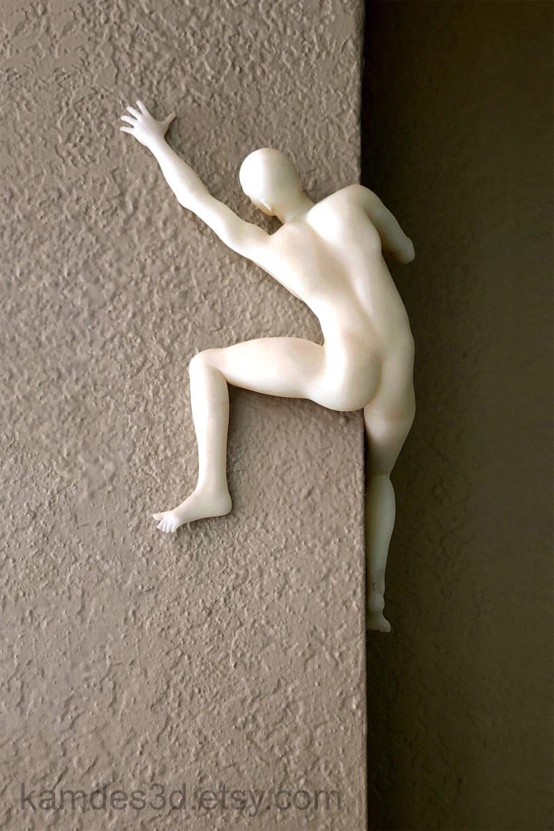 May include: A matte off-white figurine of a human figure in a climbing pose, mounted on a corner edge. The figure has one arm extended upward and one leg bent, with the body positioned to appear as if it is scaling the vertical surface of the wall.