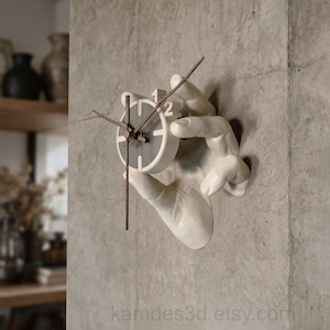 May include: A wall-mounted clock featuring a sculpted white hand holding a circular clock face. The clock face is white with a dark gray center, thin dark brown hands, and the number 2 clearly visible. The text kamdes3d.etsy.com is printed below.