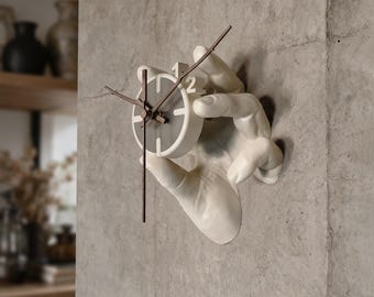 May include: A wall-mounted clock featuring a sculpted white hand holding a circular clock face. The clock face is white with a dark grey centre, thin dark hands, and the number 2 in white. The text kamdes3d.etsy.com is visible at the bottom.