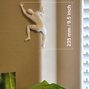 May include: A white wall-mounted sculpture of a human figure climbing a corner. The figure is depicted in a dynamic pose with arms and legs extended. Text on the image indicates a height of 9.5 inches, which corresponds to 235 mm.