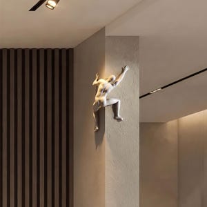 May include: A white wall-mounted sculpture of a human figure climbing around the corner of a wall. The figure is depicted in a dynamic pose with one arm reaching upward and legs bent as if scaling the surface. The text kamdes3d.etsy.com is visible.