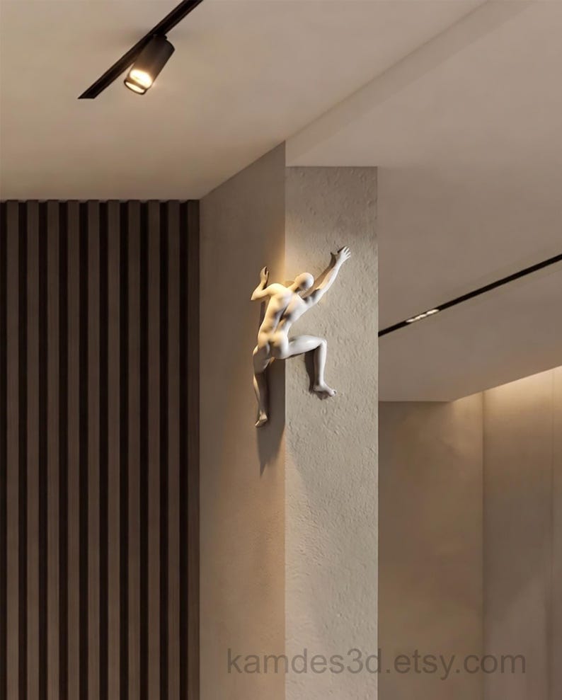 May include: A white wall-mounted sculpture of a human figure climbing around the corner of a wall. The figure is depicted in a dynamic pose with one arm reaching upward and legs bent as if scaling the surface. The text kamdes3d.etsy.com is visible.