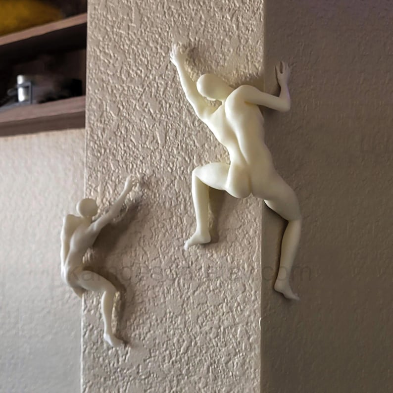 May include: Two off-white, smooth-surfaced wall sculptures depicting human figures in climbing poses. One figure is positioned on the flat surface of the wall, while the other figure is mounted on the corner edge, appearing to scale the vertical structure.
