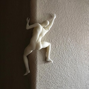 May include: A matte off-white wall sculpture depicting a human figure in a climbing pose. The figure is positioned at a corner, with one hand and one foot on one side of the wall and the other hand and foot on the adjacent side, creating a dynamic effect.