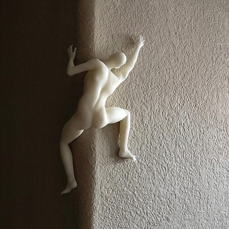 May include: A matte off-white wall sculpture depicting a human figure in a climbing pose. The figure is positioned at a corner, with one hand and one foot on one side of the wall and the other hand and foot on the adjacent side, creating a dynamic effect.
