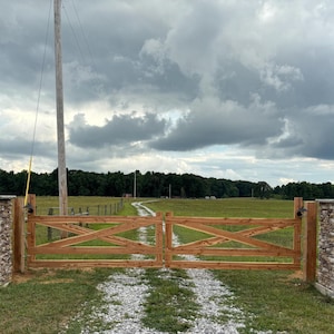 May include: A double wooden gate with a cross-brace design mounted between two stone pillars. The gate is made of light brown wood and features horizontal rails. The stone pillars are constructed from stacked, multi-colored natural stones.