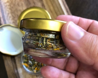 May include: A small glass jar with a gold lid filled with dried herbs and flowers. The jar is being held in a hand.