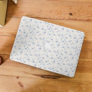 May include: A laptop case featuring a white background adorned with a blue floral pattern. The case is placed on a wooden surface. A vintage telephone and a drink are also visible. The floral design covers the entire surface of the case.