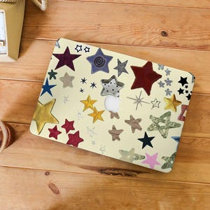 Pode incluir: Uma capa para laptop com fundo creme é coberta por várias formas de estrelas coloridas. As estrelas são em tons de vermelho, amarelo, azul, verde e preto. A capa está sobre uma superfície de madeira.