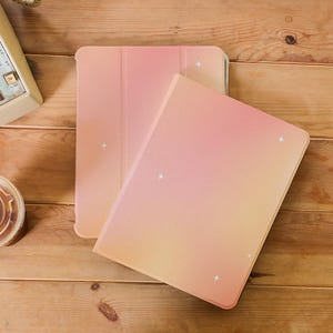 Può includere: Due custodie per tablet con un design sfumato nei toni del rosa e del giallo, decorate con piccoli accenti di stelle bianche. Le custodie sono aperte, rivelando l'interno, su una superficie di legno.