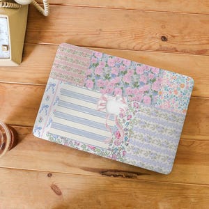 Puede incluir: Una funda rectangular para portátil con un diseño de patchwork. Presenta patrones florales y de rayas en rosa pastel, azul y lavanda. Un lazo blanco con cintas rosas está centrado en la funda. Sobre una superficie de madera.