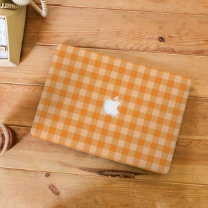 May include: An orange and cream gingham pattern laptop case with the Apple logo visible. The case is on a wooden surface, with a vintage telephone and a beverage cup in the background.