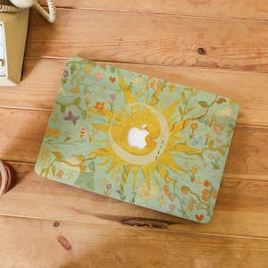 May include: A laptop case with a whimsical design featuring a yellow sun and crescent moon, surrounded by flowers, butterflies, and hearts on a light green background. The case is on a wooden surface.