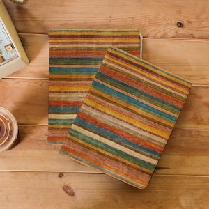 Może przedstawiać: Dwa prostokątne notatniki lub czasopisma w wielokolorowe paski. Paski obejmują odcienie pomarańczowego, żółtego, niebieskiego i brązowego. Notatniki leżą na drewnianej powierzchni, z zabytkowym telefonem i filiżanką kawy.