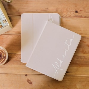 May include: A light beige tablet case with the name "Alberta" in a cursive font. The case is open, revealing the interior, and rests on a wooden surface. A landline phone and a coffee cup are visible in the background.