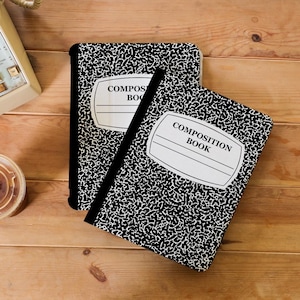 Puede incluir: Dos cuadernos de composición rectangulares con un patrón clásico de manchas negras y blancas en las portadas. Cada cuaderno tiene una etiqueta central blanca con el texto impreso COMPOSITION BOOK y líneas horizontales para escribir.