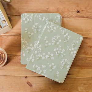 May include: Two light green tablet cases with a white floral design. The cases are open, showing the inside. They are on a wooden surface. A vintage telephone and a coffee cup are in the background.