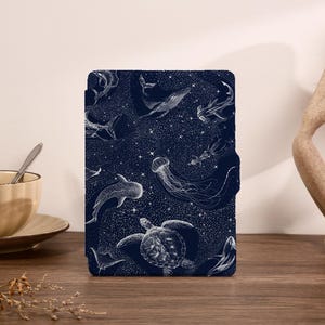 Może przedstawiać: Ciemnoniebieskie etui na tablet z białą ilustracją różnych stworzeń morskich, w tym wielorybów, rekinów i meduz, na tle gwiaździstego nieba. Etui jest prostokątne z magnetycznym zamknięciem.