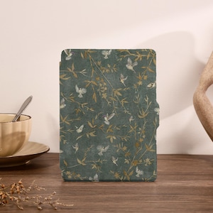 May include: A rectangular e-reader case featuring a dark teal background with a repeating pattern of gold-toned bamboo branches and small white birds in flight. The case has a smooth, matte finish and a slim profile with a small side tab.