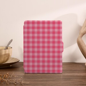 May include: A pink and light pink checkered tablet case stands upright on a wooden surface. The case is the focal point, with a teacup and saucer to the left and a decorative sculpture to the right. The case has a simple, clean design.