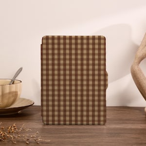 May include: A brown and tan checked tablet case stands upright on a wooden surface. A beige teacup with a spoon rests on a saucer to the left. A decorative sculpture is on the right.