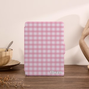May include: A pink and white gingham pattern tablet case with the name "Owen" printed in a white font. The case is standing upright on a wooden surface. A cup and saucer are visible in the background.