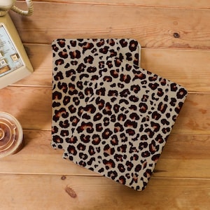 Puede incluir: Dos fundas con estampado de leopardo, posiblemente para una tableta o un cuaderno, se muestran sobre una superficie de madera. Las fundas presentan un fondo beige con manchas marrones y marrón oscuro. Las fundas son rectangulares con esquinas redondeadas.