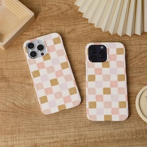 May include: Two phone cases with a pink, tan, and white chequered pattern. The cases are on a wooden surface. The phone cases have camera cutouts. The cases are designed for a mobile phone.