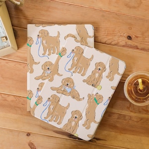 Puede incluir: Dos fundas para iPad de color crema con un patrón repetido de perros golden retriever en varias poses. Los perros están ilustrados en un estilo de dibujos animados y llevan collares azules. Algunos perros llevan correas azules en la boca.