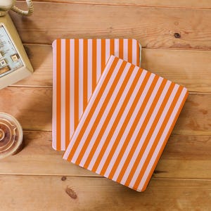 May include: Two notebooks with orange and white vertical stripes are shown on a wooden surface. The notebooks are rectangular with rounded corners. A vintage telephone and a coffee cup are in the background.