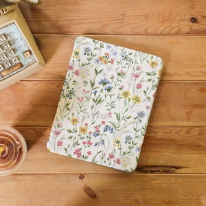 May include: A floral-patterned tablet case featuring a white background with colorful wildflowers in shades of pink, yellow, and blue. The case is on a wooden surface, with a vintage telephone and a cup of coffee nearby.