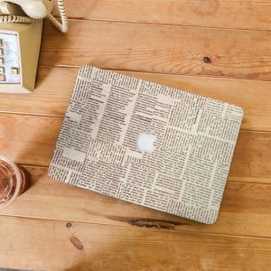 Puede incluir: Una funda para portátil con un diseño de impresión de periódico. La funda es beige con texto negro y un logotipo de Apple blanco.