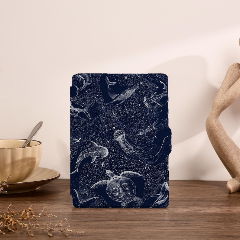 May include: A rectangular e-reader case featuring a dark navy blue background with a white line-art pattern of various sea creatures including a whale, a sea turtle, a jellyfish, and sharks, all set against a field of small white stars.