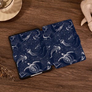 May include: A dark blue e-reader case featuring a white line art pattern of various marine life including sea turtles, whales, jellyfish, and sharks set against a background of scattered stars and celestial dots across the entire surface.