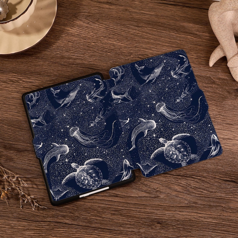 May include: A dark blue e-reader case featuring a white line art pattern of various marine life including sea turtles, whales, jellyfish, and sharks set against a background of scattered stars and celestial dots across the entire surface.