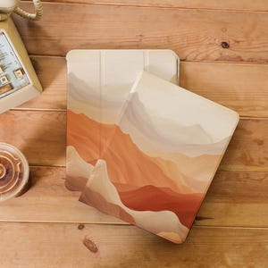 Puede incluir: Dos fundas para tablet con un diseño de paisaje de un desierto en tonos marrones, naranjas y beige. Las fundas están hechas de un material suave y texturizado.