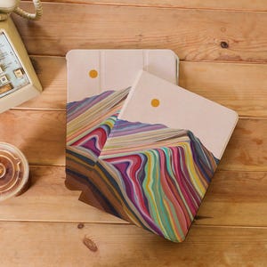 Puede incluir: Dos fundas para tablet con un diseño abstracto de paisaje de montaña colorido. Las fundas son beige con una textura sutil y tienen un sol amarillo en la esquina superior derecha.