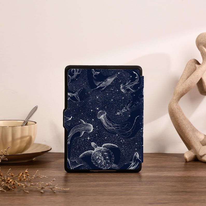 May include: A rectangular e-reader case featuring a dark navy blue background with a white line-art pattern of various sea creatures including a sea turtle, jellyfish, and whales, interspersed with small white stars across the entire surface.
