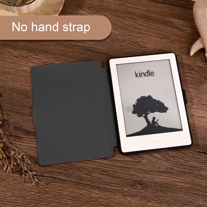May include: A white Kindle e-reader with a black frame and a dark gray protective case. The screen displays a black and white graphic of a person sitting under a tree. A text box in the upper left corner of the image states, No hand strap.
