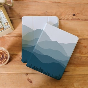 May include: Two tablet cases with a mountain range design. The cases feature a gradient of blue and gray hues, with a dark blue forest silhouette at the bottom. The cases are on a wooden surface.