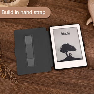 May include: A white Kindle e-reader with a black frame and a gray protective case featuring a built-in elastic hand strap. The screen displays a black and white graphic of a person sitting under a tree. The text Build in hand strap is visible.