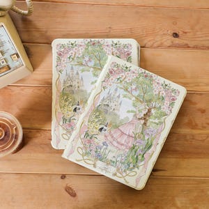 Könnte beinhalten: Zwei cremefarbene Notizbücher mit Blumendesigns. Die Umschläge zeigen eine Aquarell-Illustration eines Schlosses, eines Baumes mit rosa Blüten und einer Frau in einem rosa Kleid. Die Notizbücher haben goldene Akzente und ein Band.
