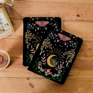 Puede incluir: Dos fundas para tablet negras con un diseño floral y celestial. El diseño presenta estrellas doradas, una luna creciente, mariposas y hongos sobre un fondo negro.