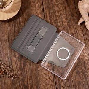 May include: A gray tablet case featuring a folding cover with an integrated elastic hand strap and a clear plastic back shell. The interior of the cover includes a magnetic ring attachment point designed for secure device mounting and alignment.