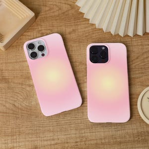 Puede incluir: Dos fundas rosas para teléfono con un diseño degradado suave. Las fundas tienen un centro amarillo claro que se desvanece en un tono rosa. Las fundas están sobre una superficie de madera.